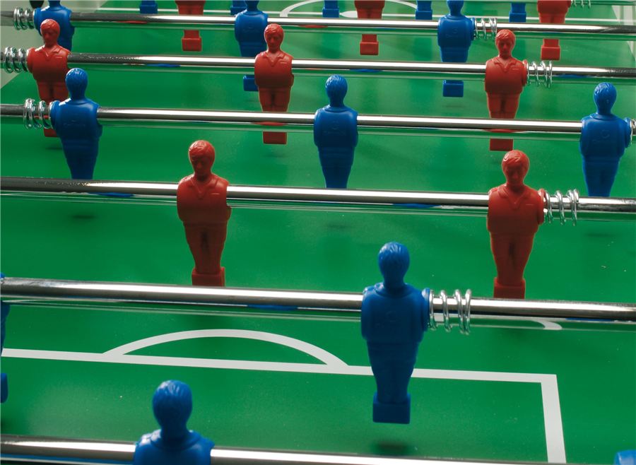 Nahaufnahme eines Kickertischs mit blauen und roten Spielfiguren auf grünem Spielfeld mit weißen Linien.