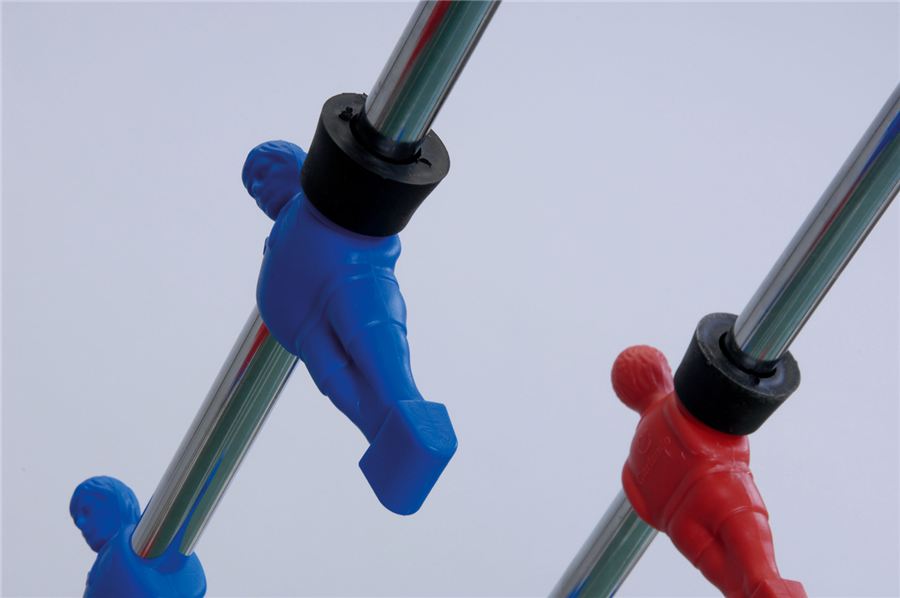 Zwei Kickerfiguren, eine blau und eine rot, sind an verchromten Stangen eines Tischkickers montiert, vor hellem Hintergrund.