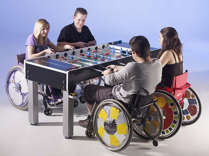 Vier Personen im Rollstuhl spielen gemeinsam Tischfußball in einem hellen Raum.
