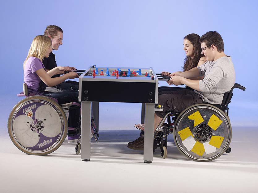 Vier Personen im Rollstuhl spielen Tischfußball an einem Tisch mit blauer und roter Spielfigur, vor blauem Hintergrund.