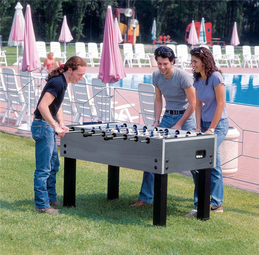 Drei junge Erwachsene spielen Tischkicker im Freien auf einer Wiese neben einem Pool mit Liegestühlen und Sonnenschirmen.