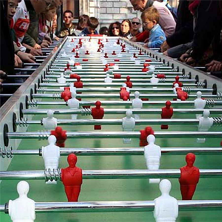 Menschenmengen versammeln sich um einen übergroßen Tischkicker mit roten und weißen Figuren, während ein Spiel im Gange ist.