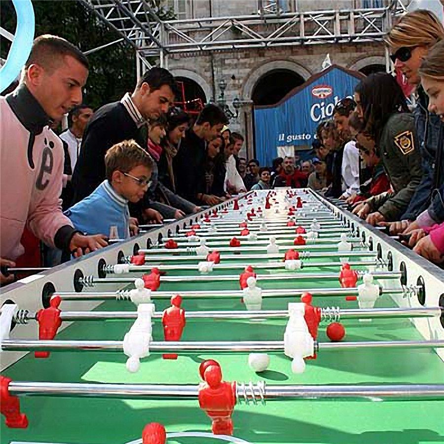 Menschenmengen spielen an einem übergroßen Tischkicker im Freien, während sie sich auf das Spiel konzentrieren.