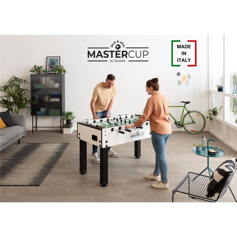 Zwei Personen spielen Tischfußball an einem weißen Kickertisch in einem modern eingerichteten, hellen Wohnzimmer mit Pflanzen und Sofa.