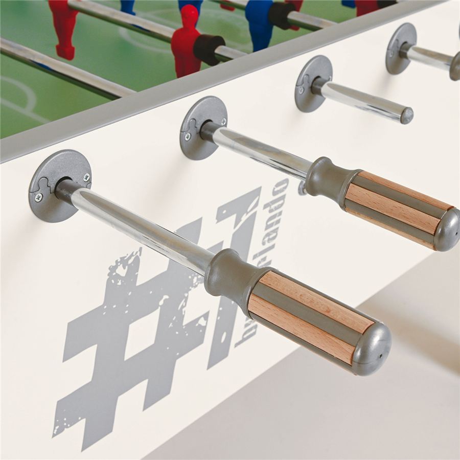 Detailaufnahme eines Kickertischs mit Holzgriffen und Metallstangen, im Hintergrund rote und blaue Spielfiguren auf grünem Spielfeld.