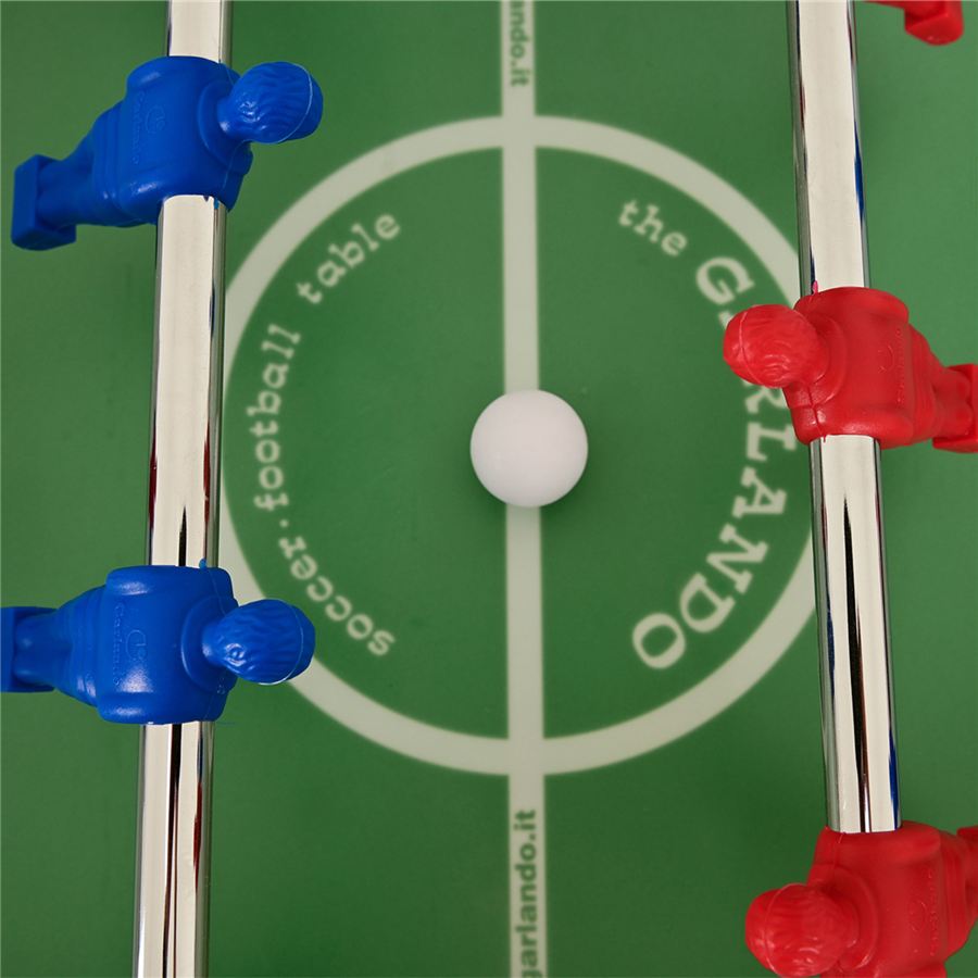 Tischfußball-Spielfeld mit blauen und roten Spielfiguren an Metallstangen und einem weißen Ball auf dem Anstoßpunkt.